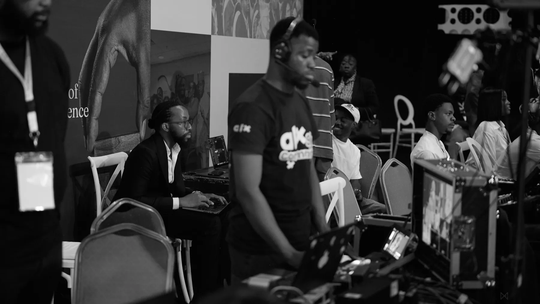 David ’Tobi Oyawoye at a laptop beside the stage, the crew at work around him