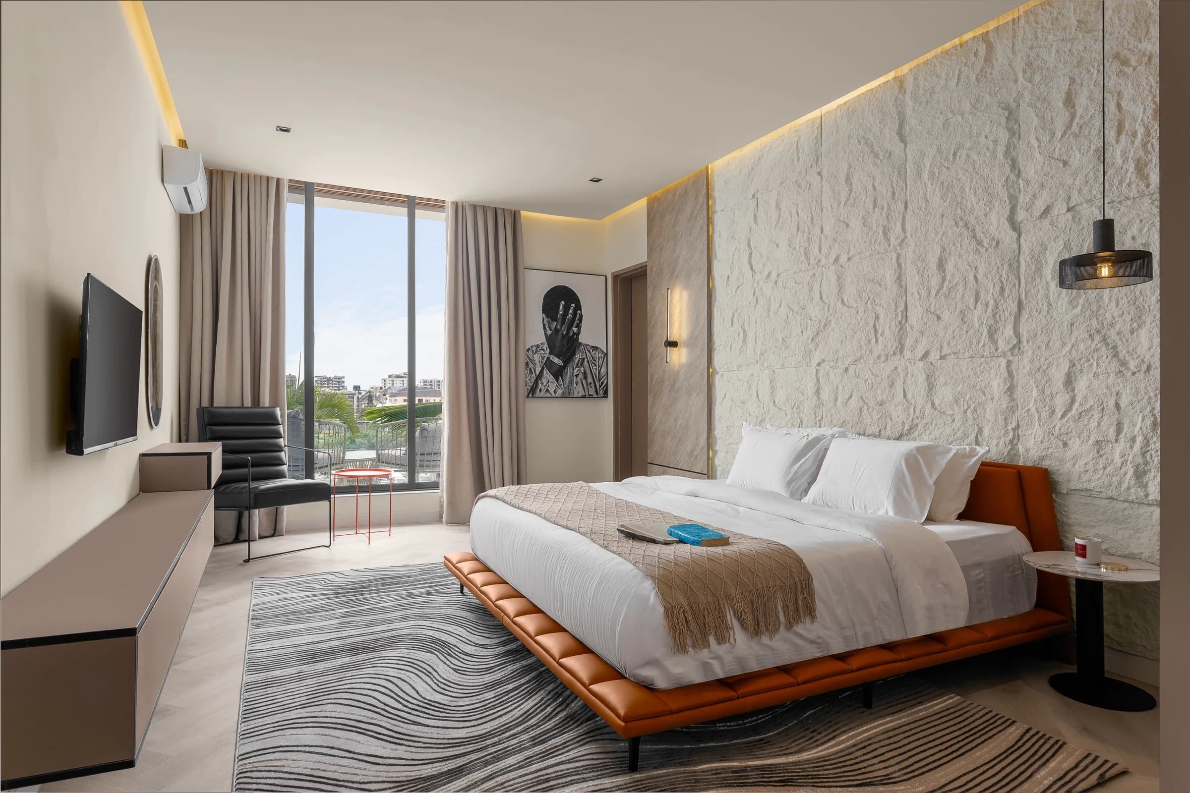 A Nook bedroom: a low tan leather bed against a white stone wall, a window over Lagos